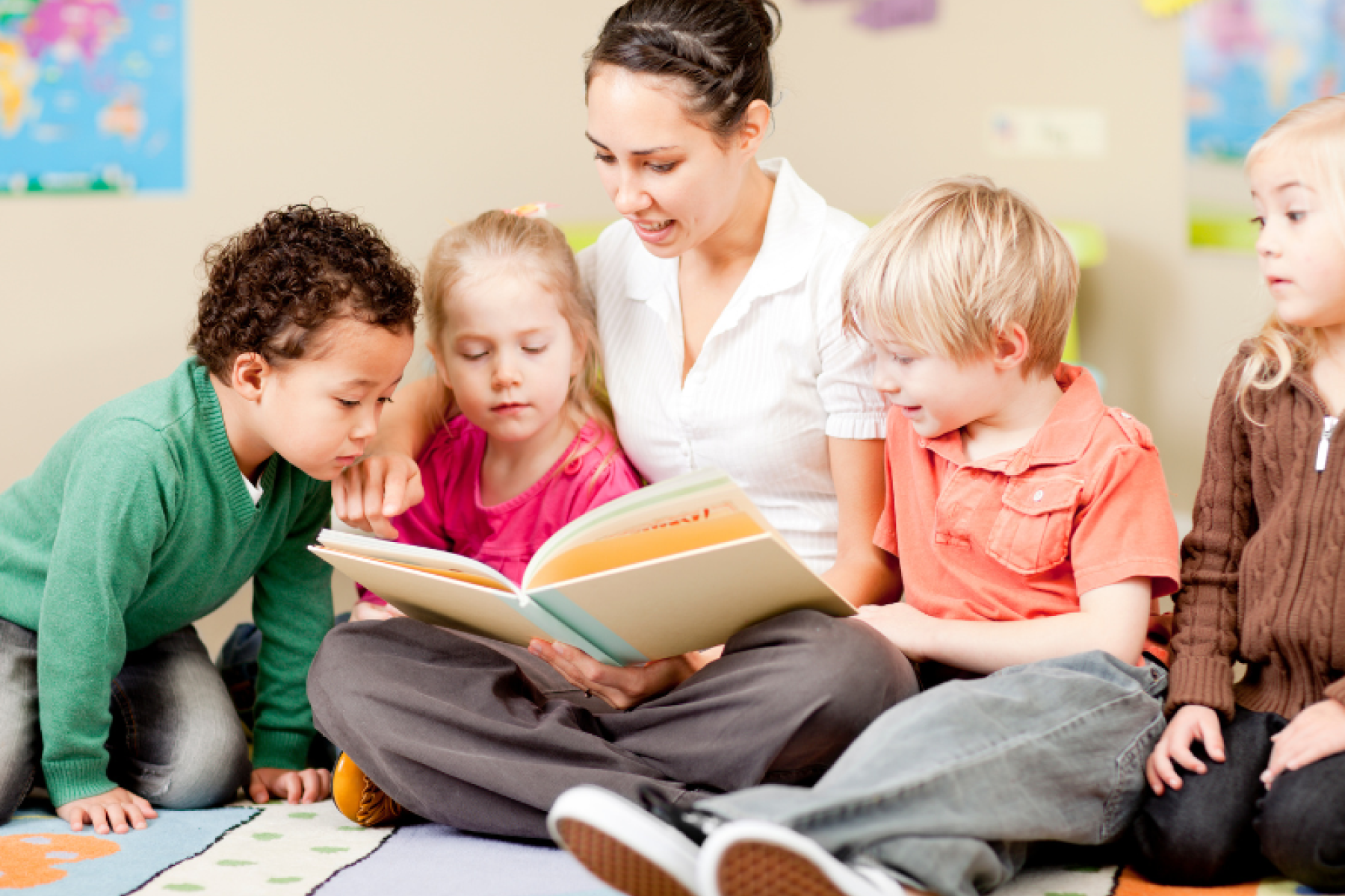 Eine Frau sitzt mit eine Gruppe von Kindergartenkindern auf dem Boden und liest ein Buch.
