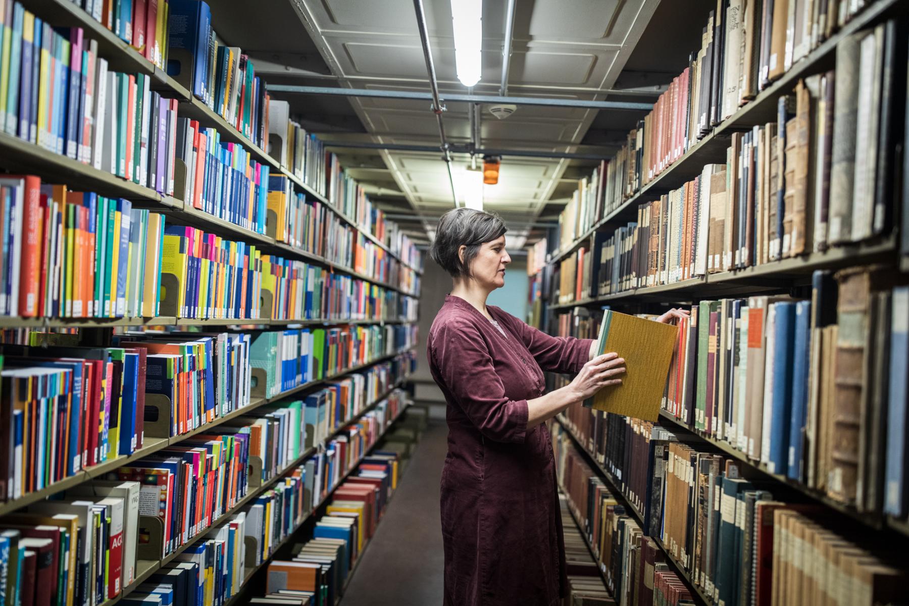Ein Frau steht zwischen zwei symetrischen Bücherregalreihen udn stellt ein Buch zurück ins Regal