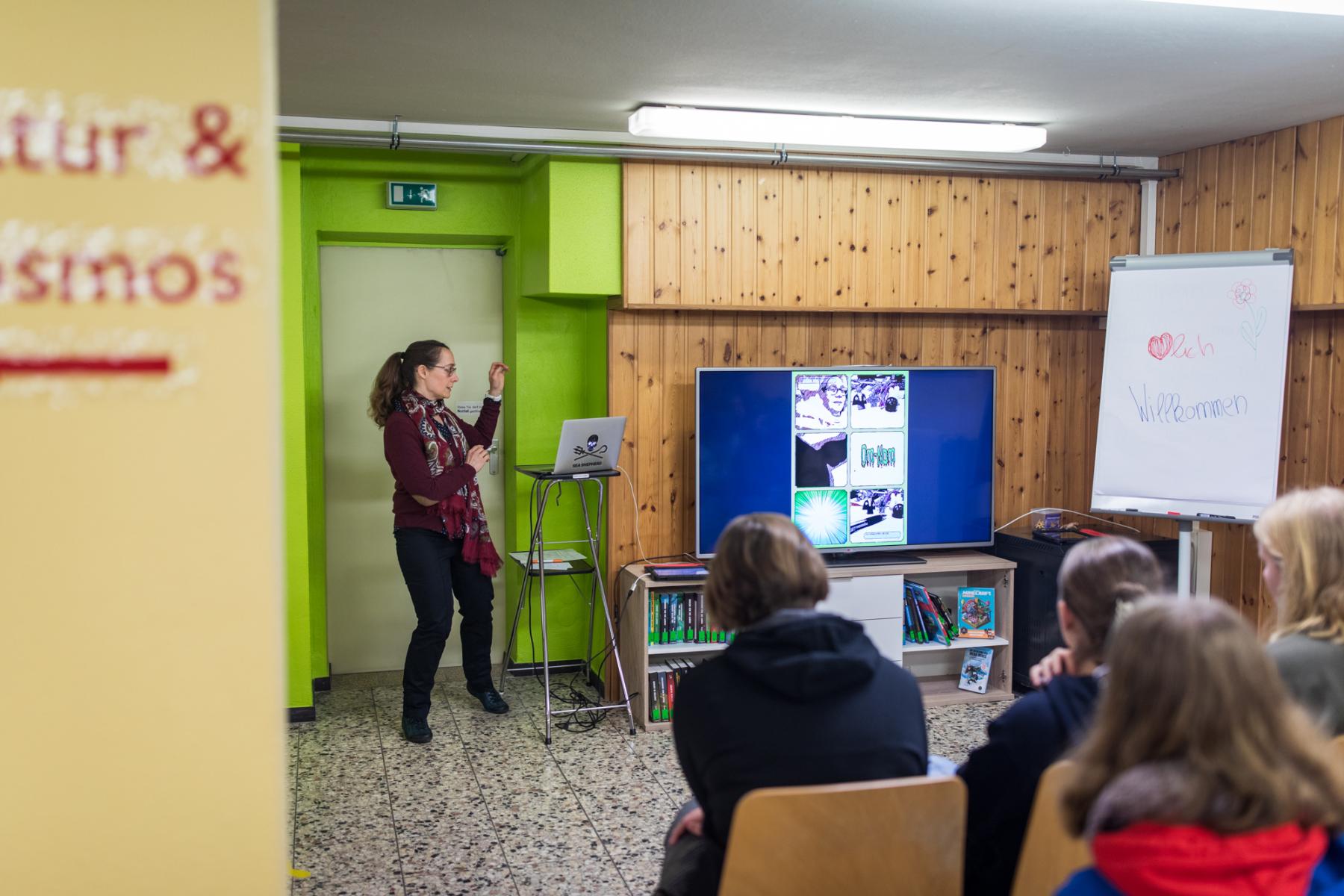 Bibliothekarin steht vor einer Gruppe mit Schülerinnen und zeigt etwas am Monitor.