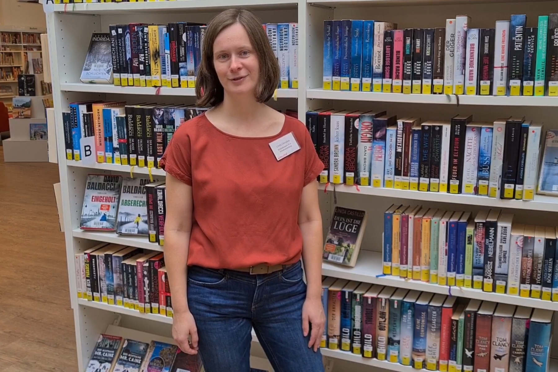 Bibliothekarin in Jeans und rotem Hemd steht vor einem bunten Bücherregal.