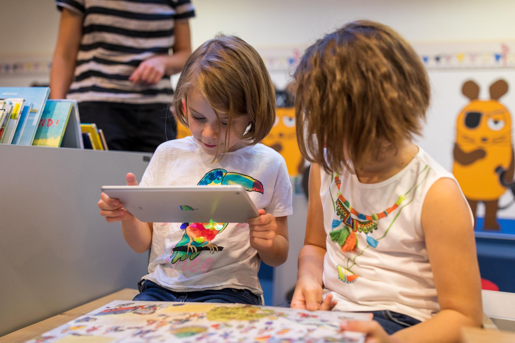 Zwei junge Mädchen sitzen über ein Buch gebeugt, ein Tablet in der Hand. Im Hintergund steht die Bibliothekarin. 