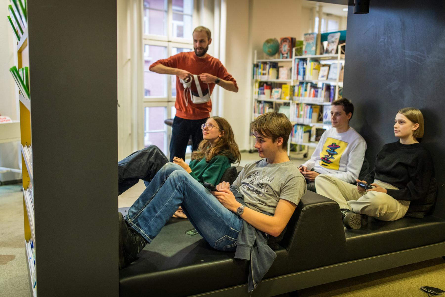 4 Jugendliche sitzen in einem Sitzgestell und spielen Konsolenspiele. Der Medienpädagoge steht dahinter und schaut zu.