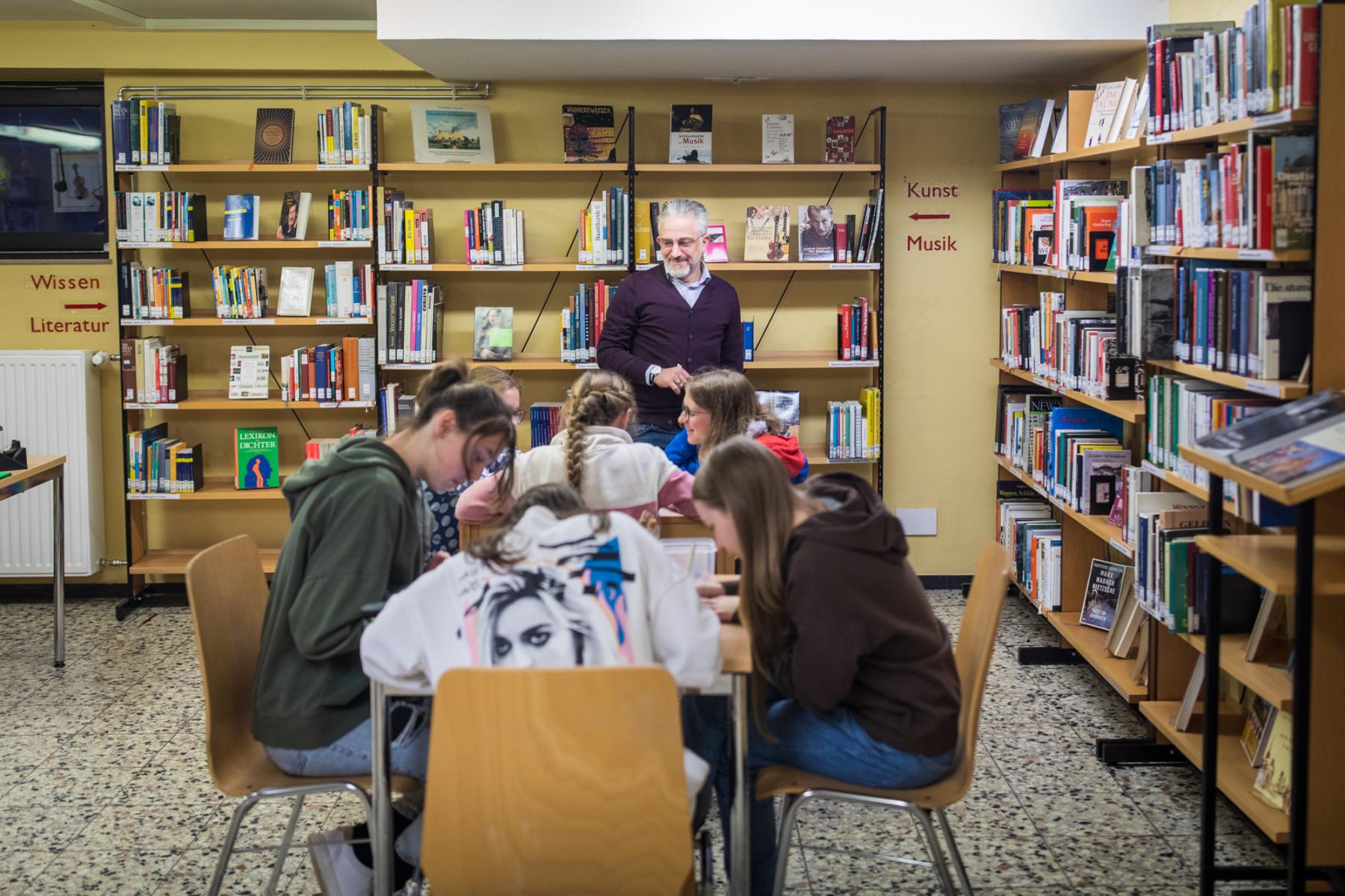 Ein Gruppe Schüler sitzt an Tischen in der Bibliothek und arbeiten konzentriert an etwas. Im Hintergrund stehen ein Lehrer und Bücherregale