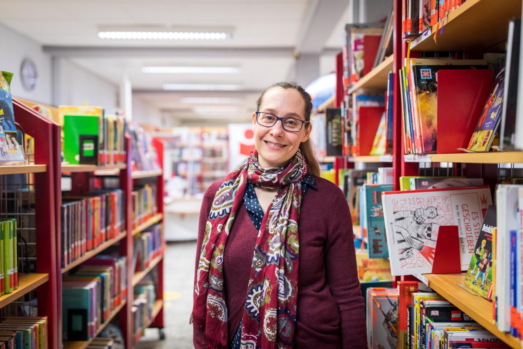 Eine Frau steht zwischen Bücherregalen in der Bibliothek und lächelt in die Kamera.