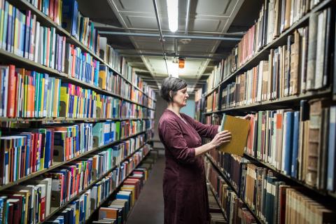 Ein Frau steht zwischen zwei symetrischen Bücherregalreihen und stellt ein Buch zurück ins Regal.