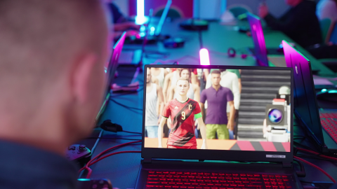 Man sieht einen Tisch mit Computern in einem abgedunkelten Raum. Frontal ist ein Laptop mit einem Videospiel zu sehen, verschwommen im Vordergund der Gamer.