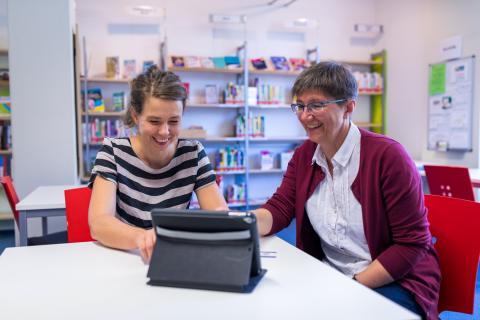 Zwei Frauen sitzen in der Bibliothek gemeinsam an einem aufgestellten Tablet und lachen.