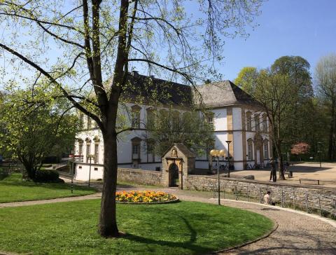 Freistehendes weißes Gebäude der Stadtbibliothek Paderborn umgeben von einer flachen Mauer, Bäumen und Wiese.