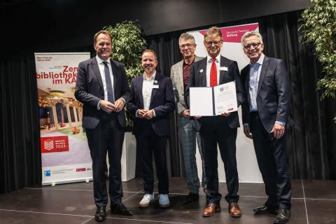 5 Männer stehen auf der Bühne, einer hält die Urkunde zur "Bibliothek des Jahres"
