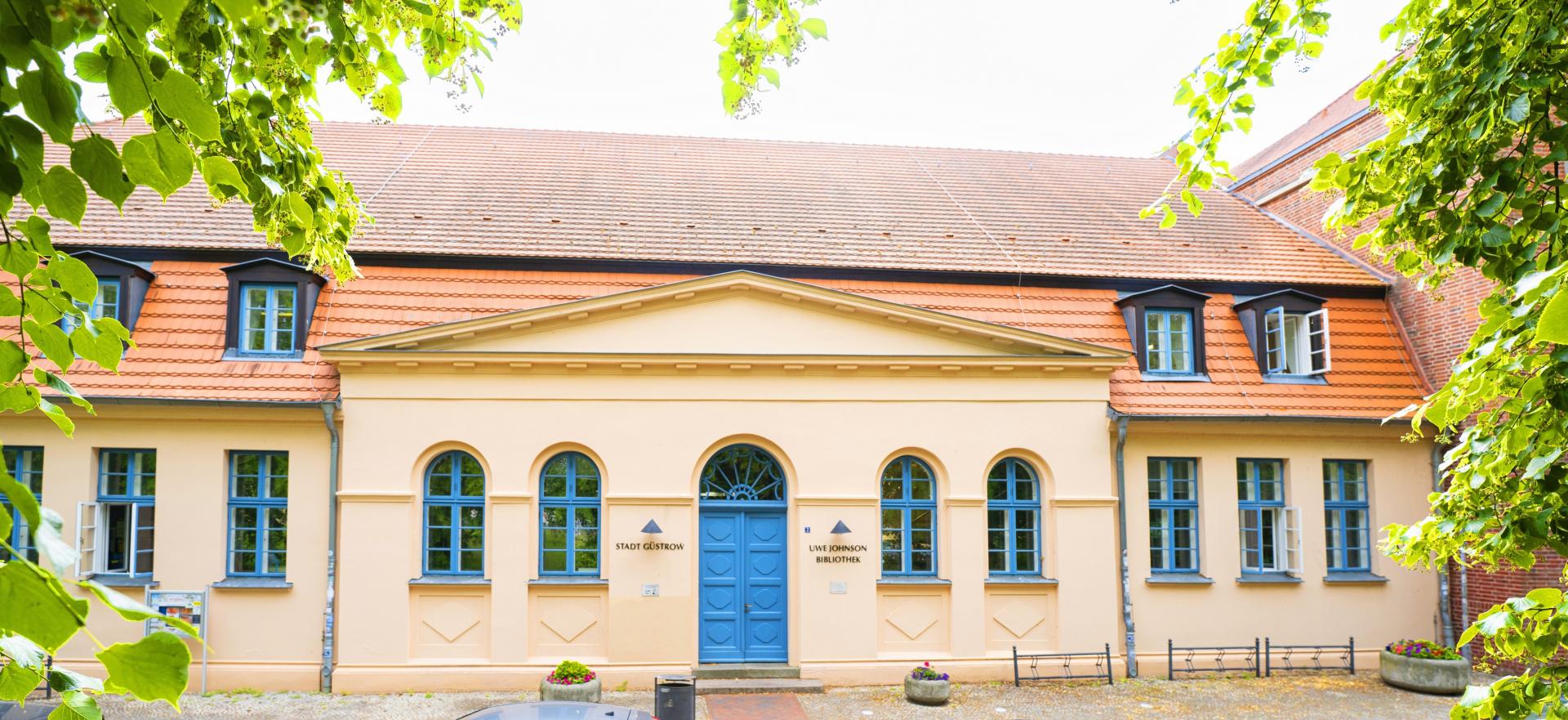 Außenansicht der okkerfarbenen Uwe Johnson-Bibliothek in Güstrow mit einer blauen Eingangstür und Bäumen links und rechts.
