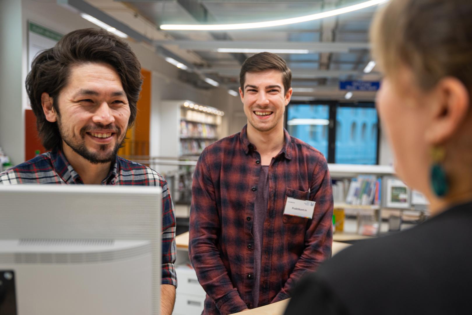 Zwei männliche Bibliotheksmitarbeiter stehen in der Bibliothek und sprechen mit einer Frau.