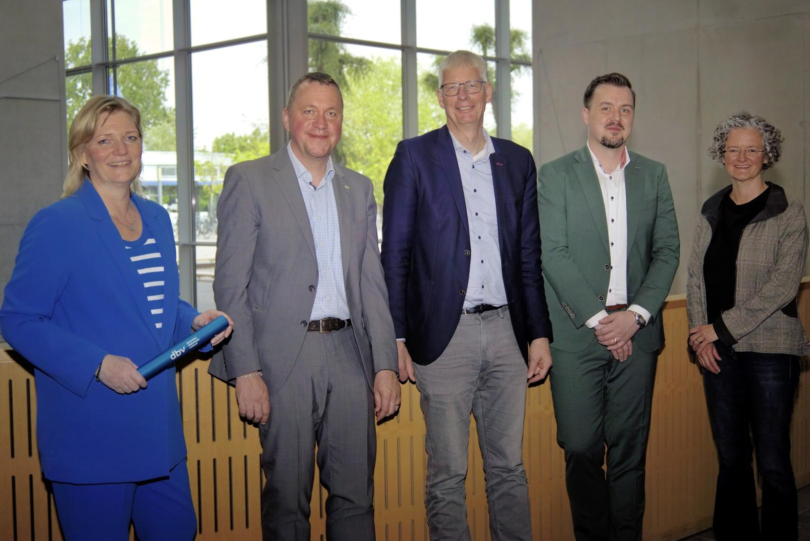 Vier Personen stehen vor einer Balustrade und lächeln in die Kamera. Die Frau ganz links hält einen Staffelstab in der Hand.