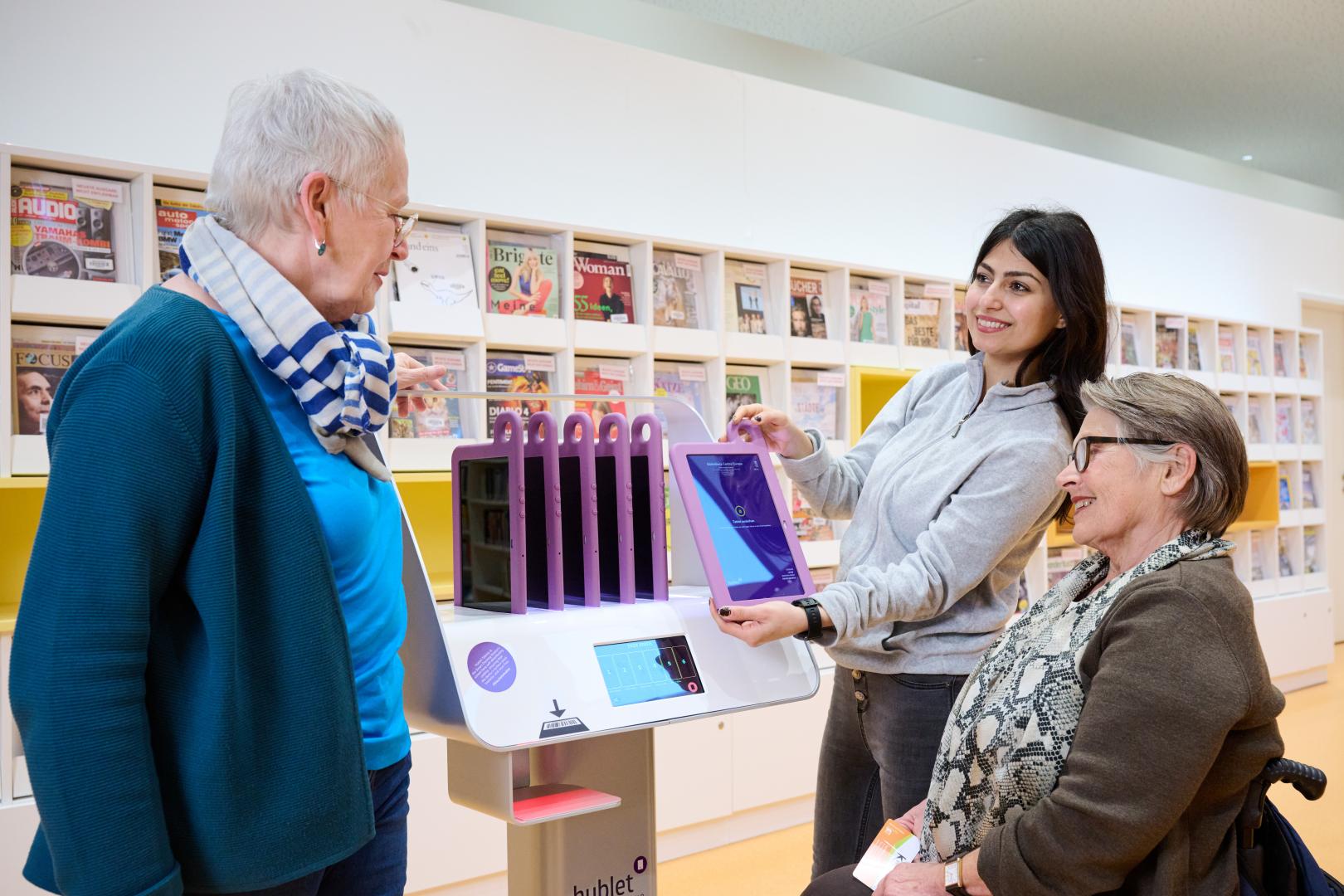 Drei Personen sind in der Bibliothek, wovon eine den anderen Tablets zeigt.