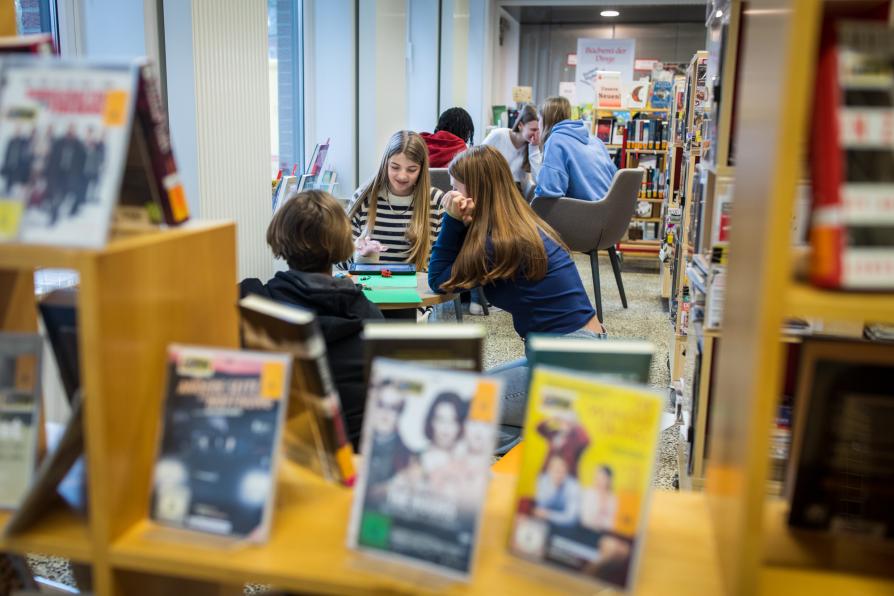 Jugendliche sitzen an kleinen Tischen in der Bibliothek und arbeiten an einer Sache. 