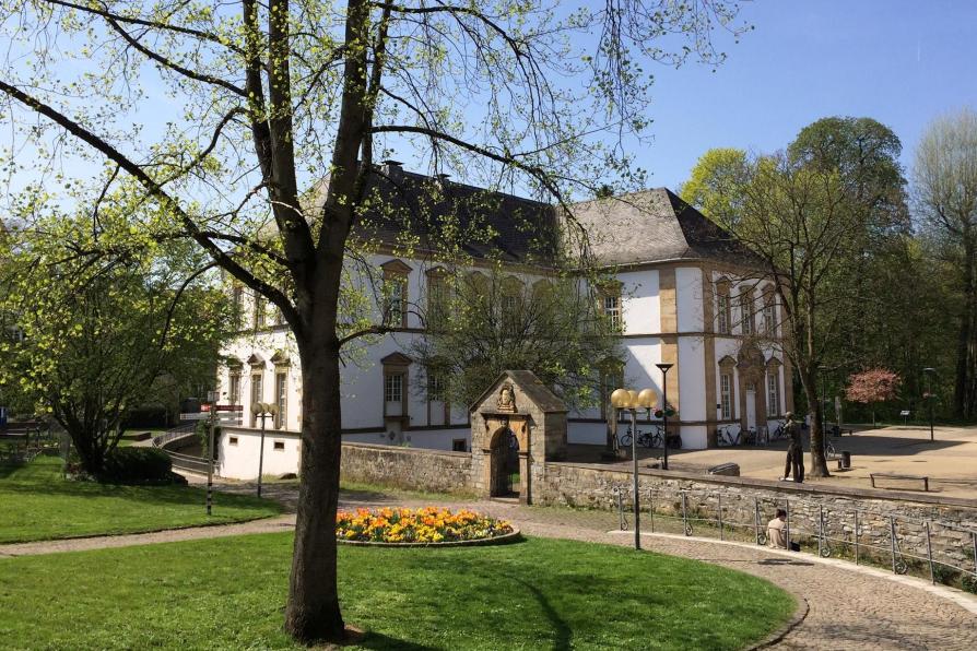 Freistehendes weißes Gebäude der Stadtbibliothek Paderborn umgeben von einer flachen Mauer, Bäumen und Wiese.