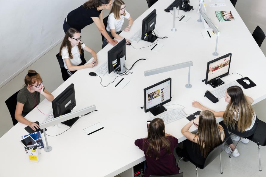 Studentinnen sitzen an Computern um einen Tisch. Eine Bibliothekarin gibt Auskunft.