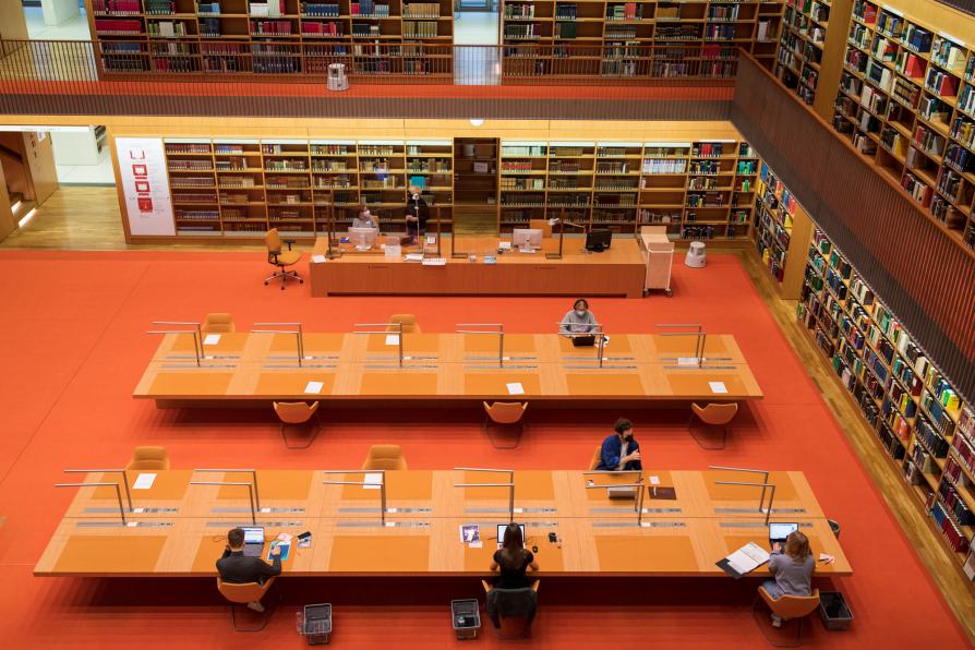 Studentinnen sitzen weit verteilt an großen Holztischen im Lesesaal der Stabi unter den Linden. Drumherum Bücherregale, darunter roter Teppichboden.