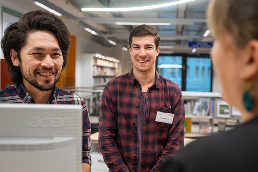 Zwei junge Männer stehen in der Bibliothek und sprechen mit einer Frau. Sie tragen Namensschilder.