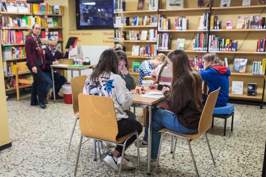 Schülerinnen sitzen in der Bibliothek an Tischen und bearbeiten Themen. Im Hintergrund sind der Lehrer und die Bibliothekarin zu sehen.