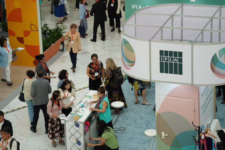 Blick in eine Messehalle mit einem Stand der IFLA. Viele Menschen gehen und stehen zwischen den Ständen.