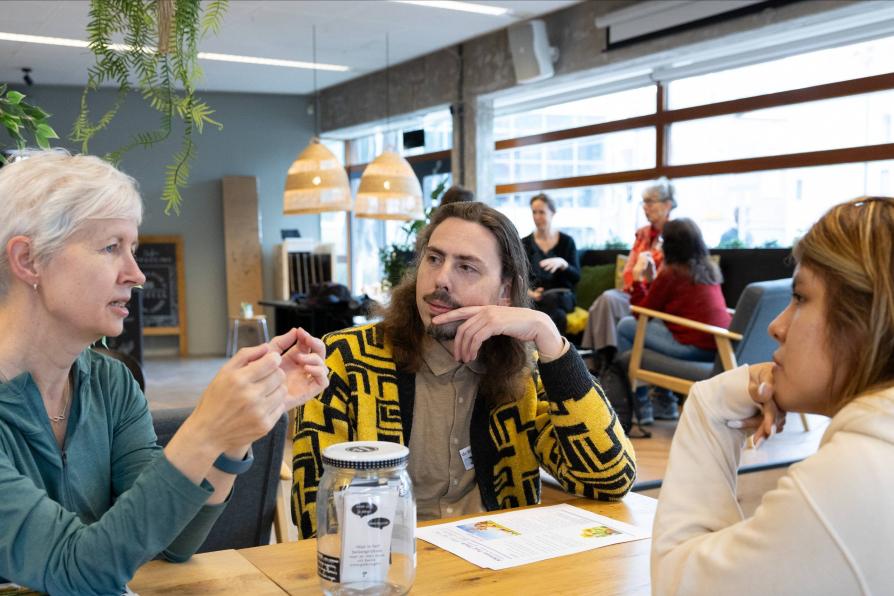 Drei Personen sitzen an einem Tisch und sprechen miteinander. Im Hintergrund sitzt eine weitere Gruppe.