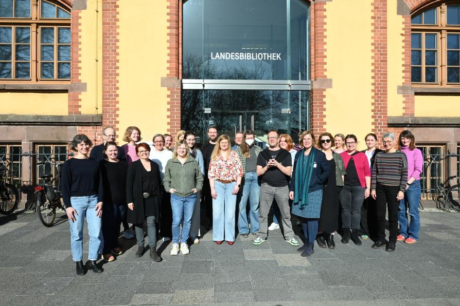 Gruppenfoto: Verschiedene Personen stehen vor einem Gebäude und lächeln in die Kamera. 