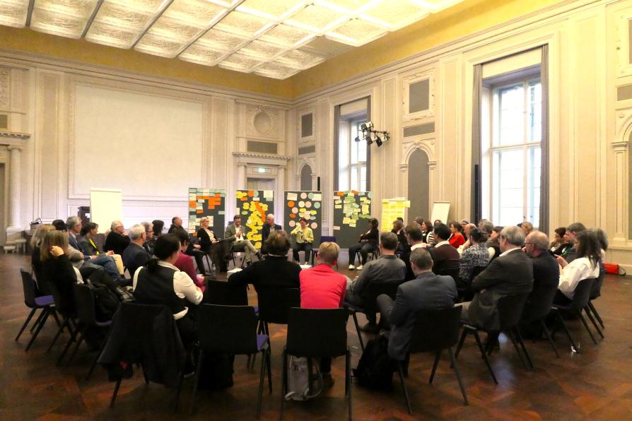 Personen sitzen im Stuhlkreis zusammen in einem Workshop, im Hintergrund sind Pinnwände mit Karten zu sehen.