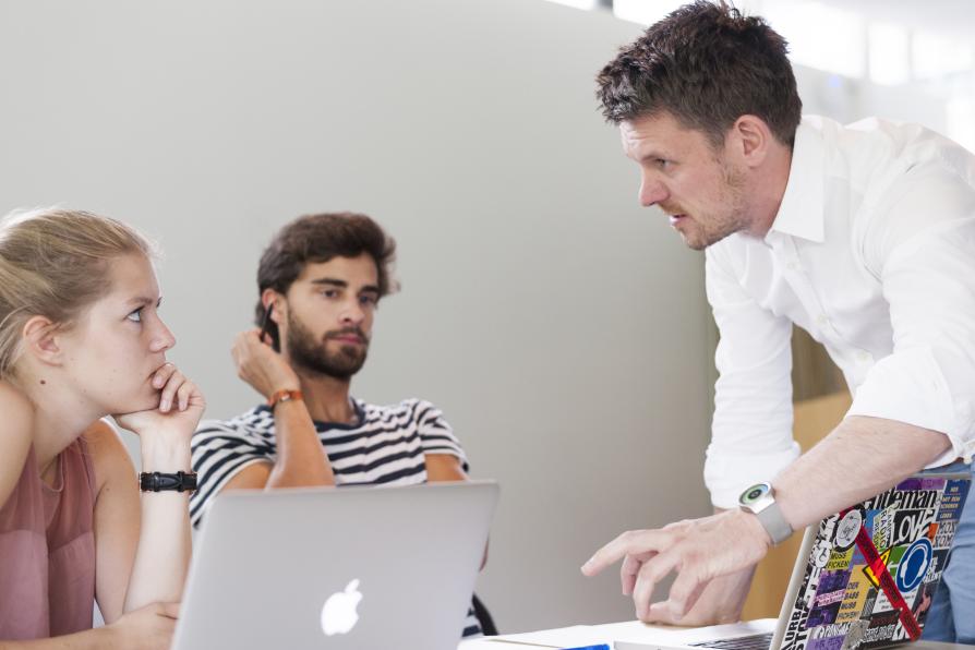 Ein Mann spricht mit einem Studenten und einer Studentin, beide haben ihre Laptops aufgeklappt vor sich stehen.